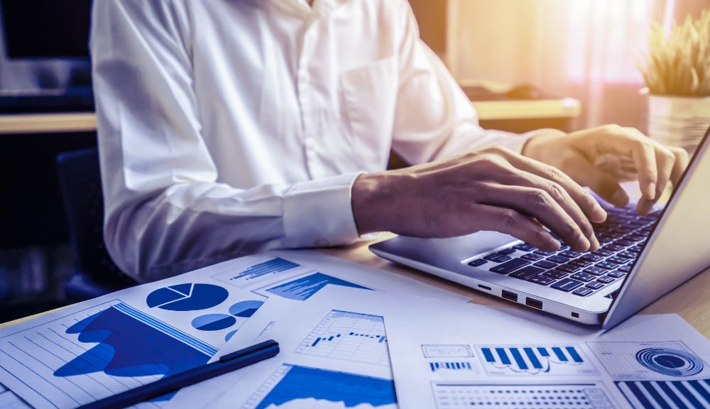 Person typing on laptop with graphs nearby.